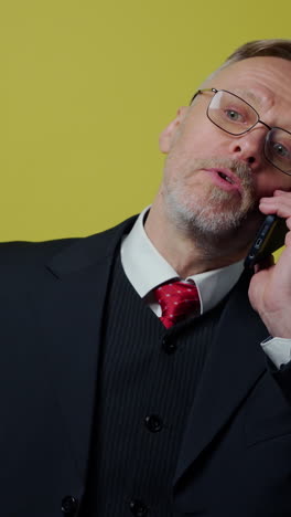 Nervous middle aged businessman with a cellphone. Mature man in dark suit talking on the phone and giving instructions to somebody on yellow background. Vertical video