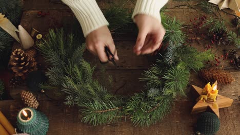 making christmas wreath. top view of woman hands making christmas wreath with fir tree and red berries on rustic wooden table with candles and pine cones. holiday advent. merry christmas