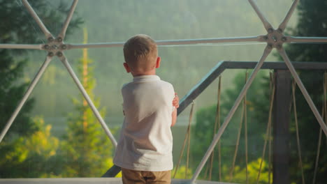 child stands in glamping tent looking at lake in distance. pensive kid in white shirt contemplates nature backside view on blurred background. calm boy rests alone at dawn