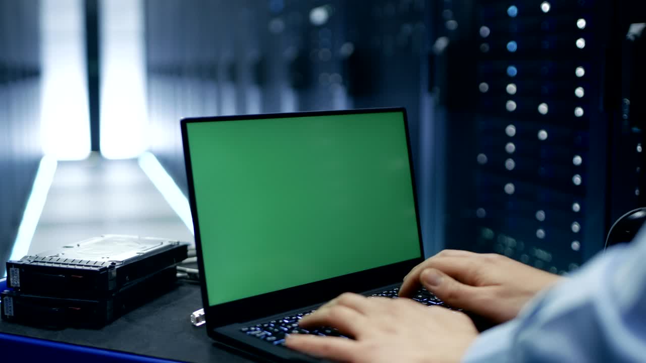 el técnico está trabajando en una computadora portátil de pantalla verde en un gran centro de datos con filas de bastidores de servidores.