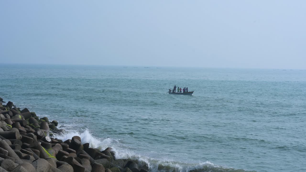 Super wide static shot of the vast Puducherry sea in early morning light, with a tiny fishing boat in the distance. Rocky shore in the left foreground and a warm sunlit skyline form the backdrop