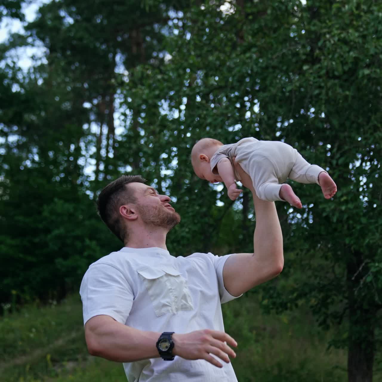 Uomo caucasico muscoloso che tiene il suo neonato sopra la testa con una mano. Papà felice e sorridente che intrattiene suo figlio. Sfondo naturale