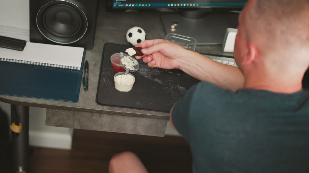 aerial view of person dipping dark snack into creamy white sauce while holding it over organized workspace with notebook, pen, soccer ball, computer monitor and keyboard visible on modern desk