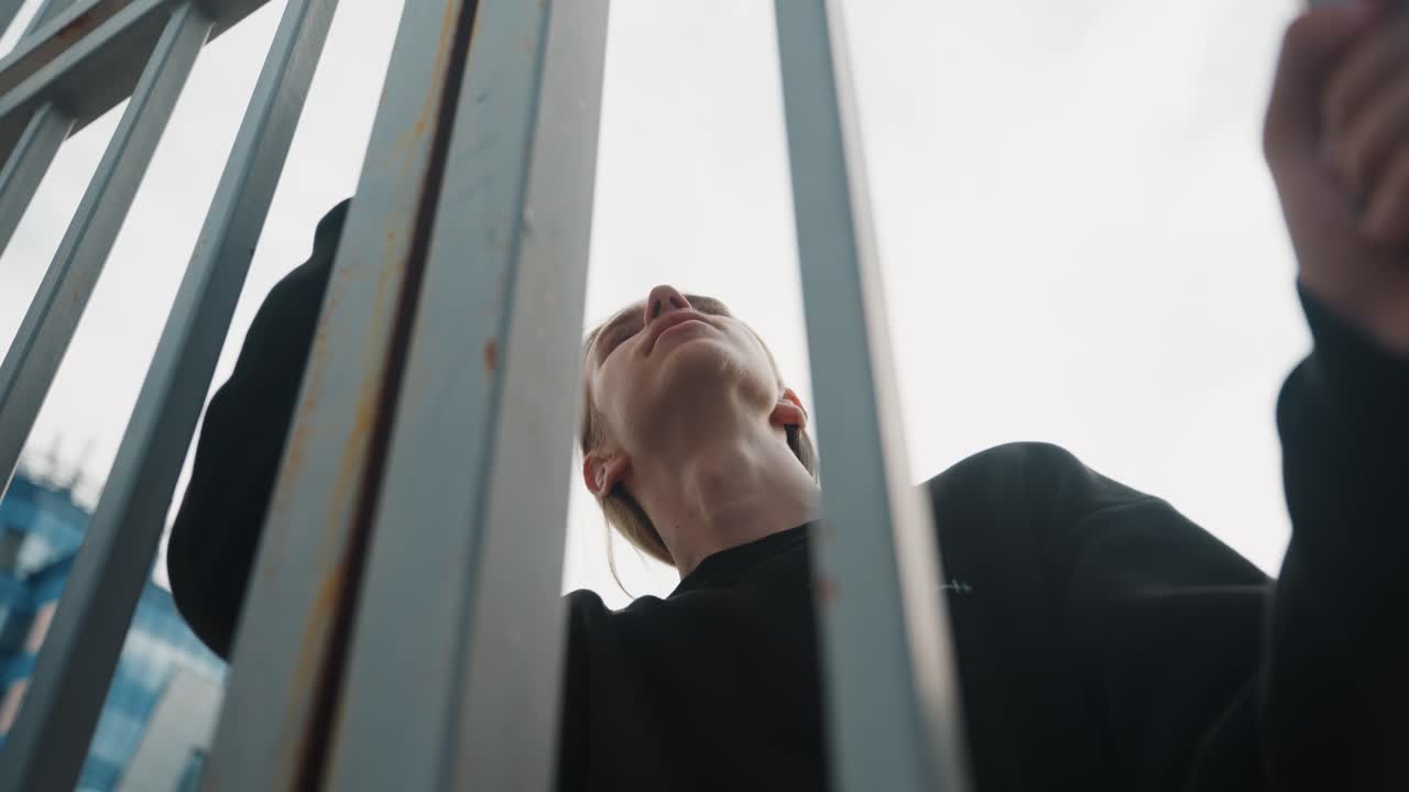 Low angle man behind metal gate looking upward, dramatic urban moment with overcast sky, tense awaiting scene suggesting access attempt or delivery arrival, bold composition with vertical rails