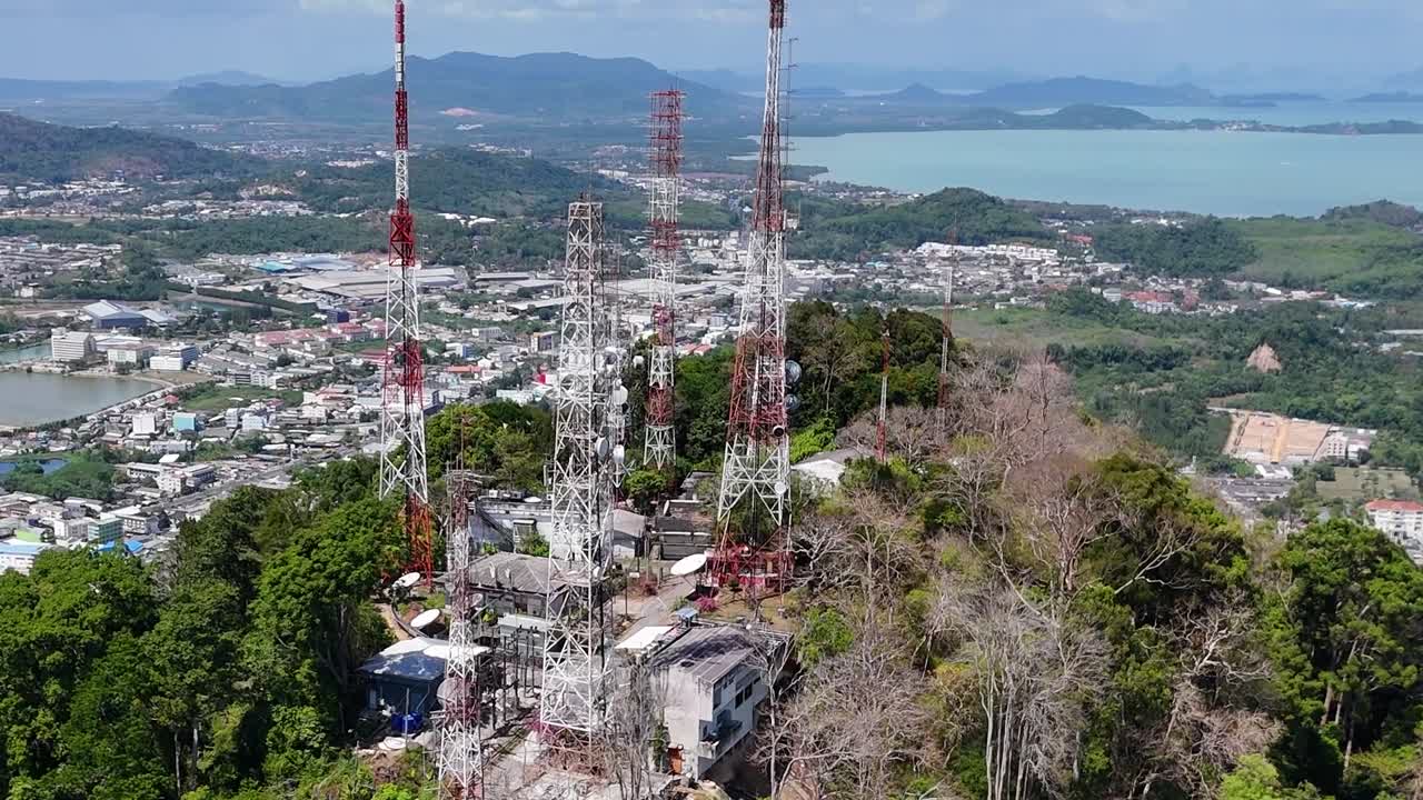 Aerial view of telecommunication towers surrounded by greenery, with a cityscape and distant waters in the background.