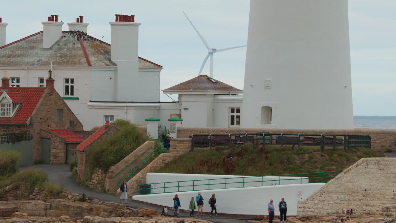 Camera pans right across St Mary’s Lighthouse and adjacent cottages, revealing visitors walking along the coastal path under overcast daylight with wind turbines in background