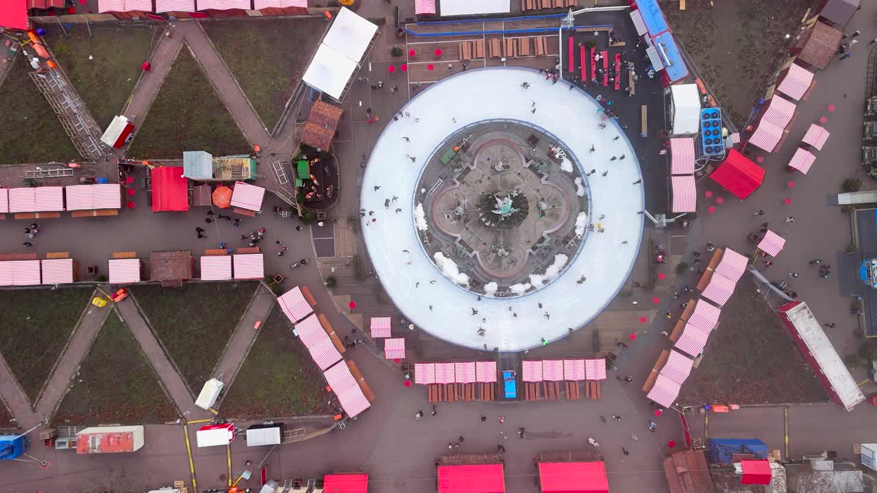 A lively winter market features people skating on a large ice rink, surrounded by cozy stalls selling food and holiday goods. The atmosphere is joyful and festive