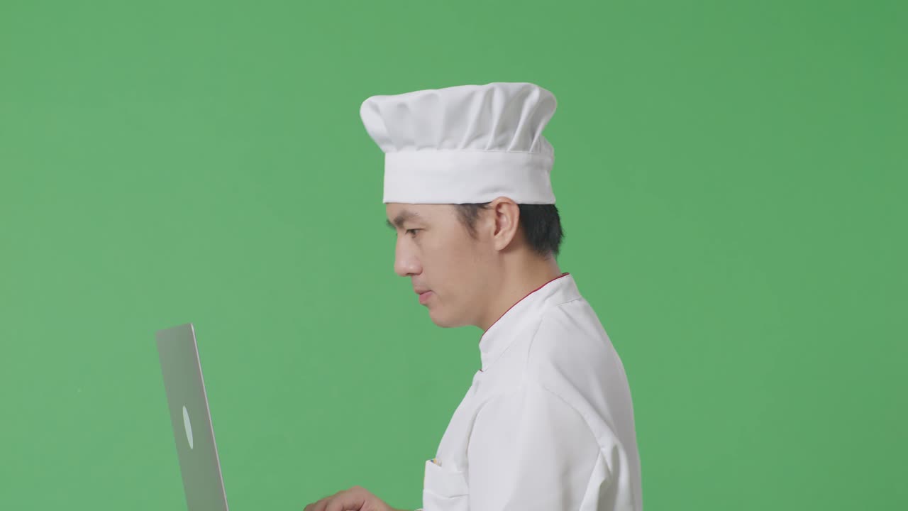 Vista laterale di uno chef asiatico che usa un laptop mentre cammina nello studio con sfondo green screen