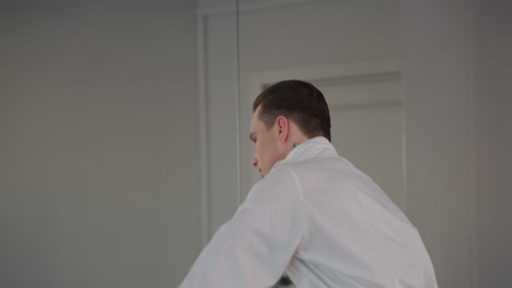 young man adjusts collar and walks toward mirror revealing neck tattoo while styling white shirt in bright modern room with soft natural light and minimalist background