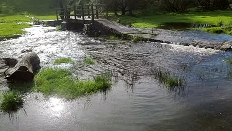 Ráfaga-Desbordante-De-Inundaciones-En-La-Orilla-Del-Río,-Soleada-Y-Pacífica-Pradera-Del-Norte-De-Gales-Bajo-El-Cruce-Del-Puente-De-Madera-Sumergido