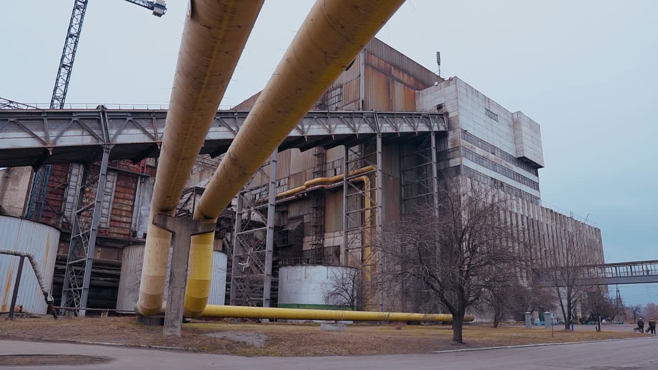 Large yellow iron pipeline leading to an old dirty rusty concrete building of a Soviet coal-fired power plant. Plant area with tanks