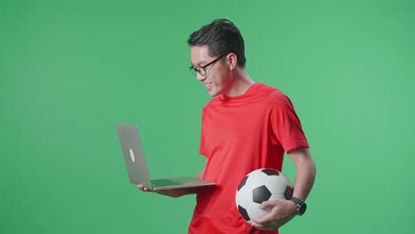 vista lateral de un hombre asiático con una pelota viendo un partido de fútbol en una computadora portátil en el estudio de pantalla verde