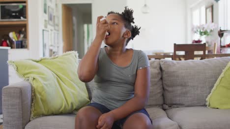 african american girl sitting on sofa in living room using inhaler