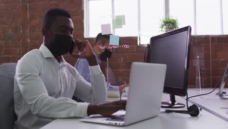 African-american-businessman-wearing-mask-sitting-on-desk-using-laptop-talking-on-smartphone