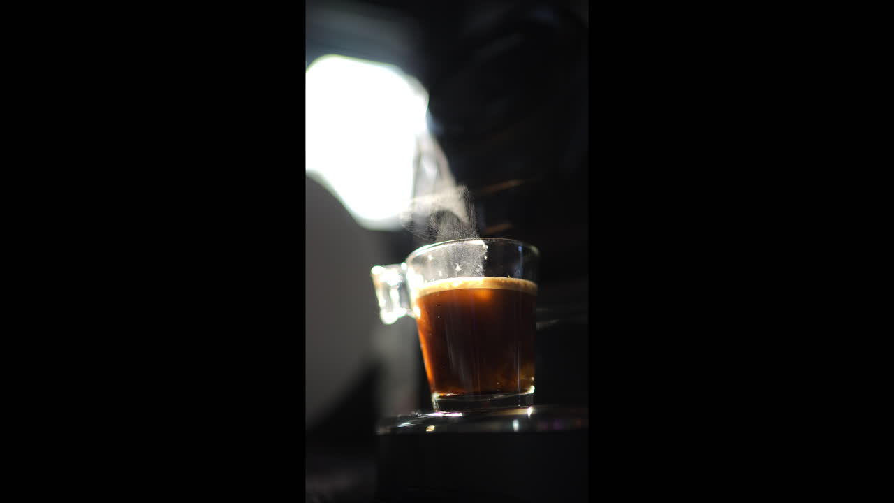 Coffee machine pouring espresso coffee into transparent glass cup on dark background, illuminated by a light beam, with steam rising from the cup