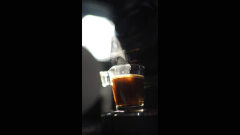 coffee machine pouring espresso coffee into transparent glass cup on dark background, illuminated by a light beam, with steam rising from the cup