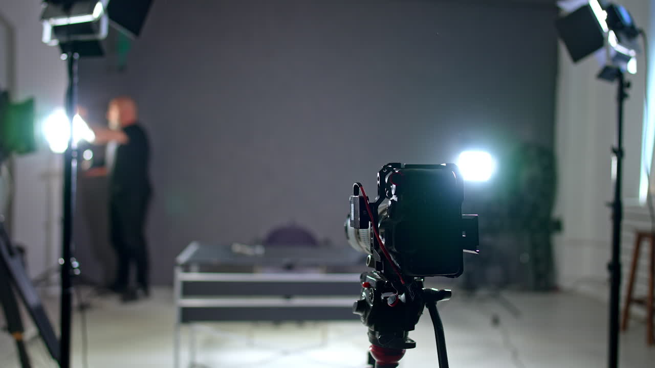 A camera on the tripod stands set in the studio. Man wearing black sets the light at blurred backdrop and then approaches camera.