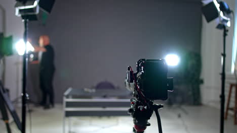 a camera on the tripod stands set in the studio. man wearing black sets the light at blurred backdrop and then approaches camera.
