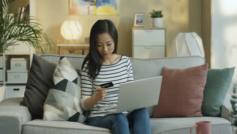 young woman wearing a striped blouse with laptop on laps and shopping online with credit card sitting on sofa in living room