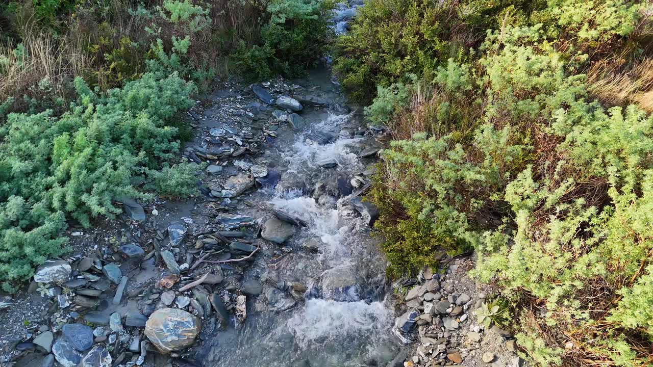 Drone footage captures a glacial stream flowing through rocky terrain surrounded by vibrant greenery in natural lighting