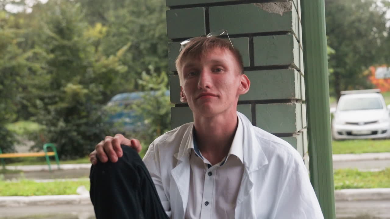 Close view of young scientific researcher in white shirt and coat sitting with relaxed posture, smiling warmly outdoors near wall, with glasses resting on head, reflecting casual human expression