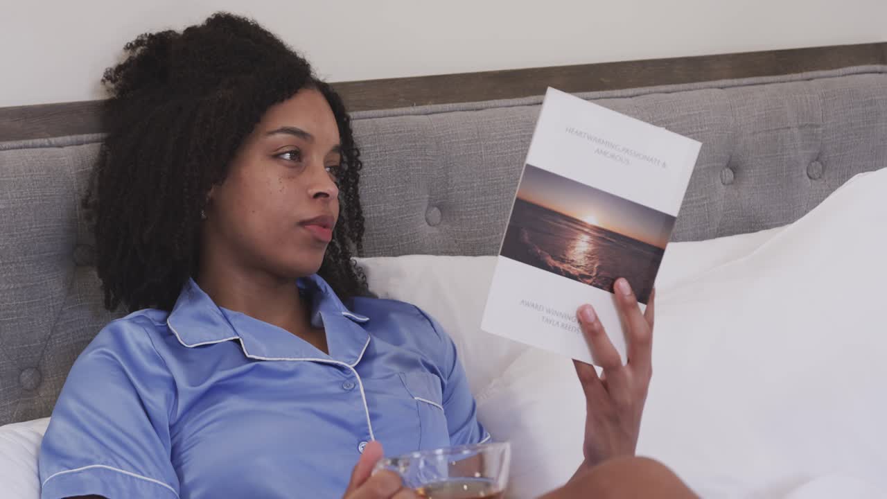 African American woman in pajamas reading book with tea in bed, camera pulling back, unwinding