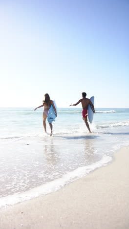 vertical video: running into ocean, couple holding surfboards, enjoying beach day together