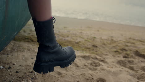 pierna de mujer con bota negra colgando de un viejo barco de pie playa de arena cerca.