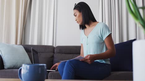 Mixed-race-gender-fluid-person-sitting-on-couch-working-from-home-using-a-laptop