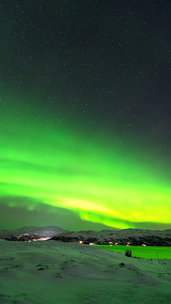The Northern Lights dance over Kvaloyvagen Norwegian town with vibrant green hues illuminating the night sky as cars rush below, vertical static