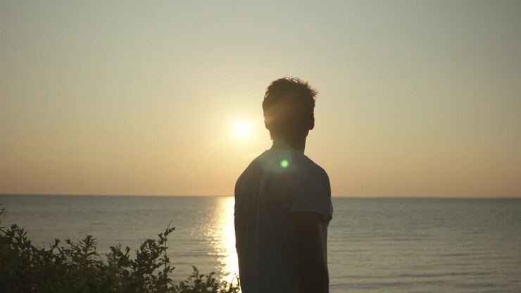 Una toma cinematográfica en cámara lenta de un hombre musculoso y en forma poniéndose AirPods y auriculares, mirando el atardecer dorado junto al océano.
