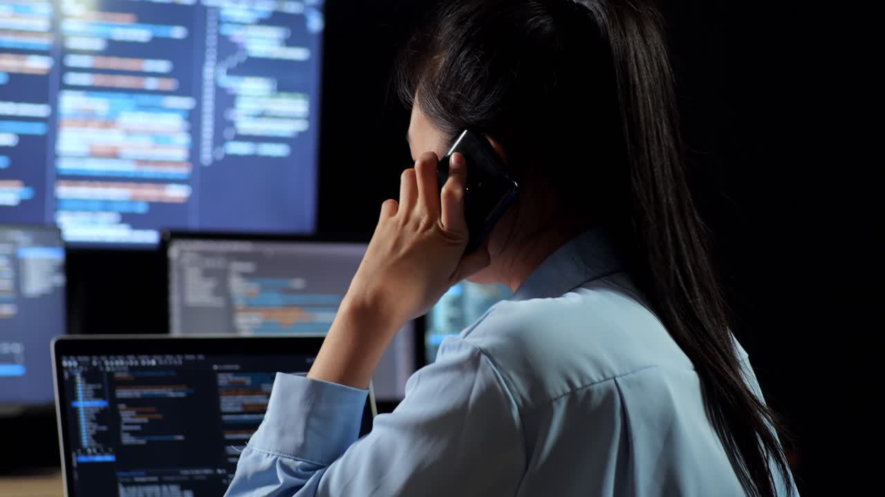 Close Up Back View Of Asian Female Programmer Talking On Smartphone While Writing Code By A Laptop Using Multiple Monitors Showing Database On Terminal Window Desktops In The Office