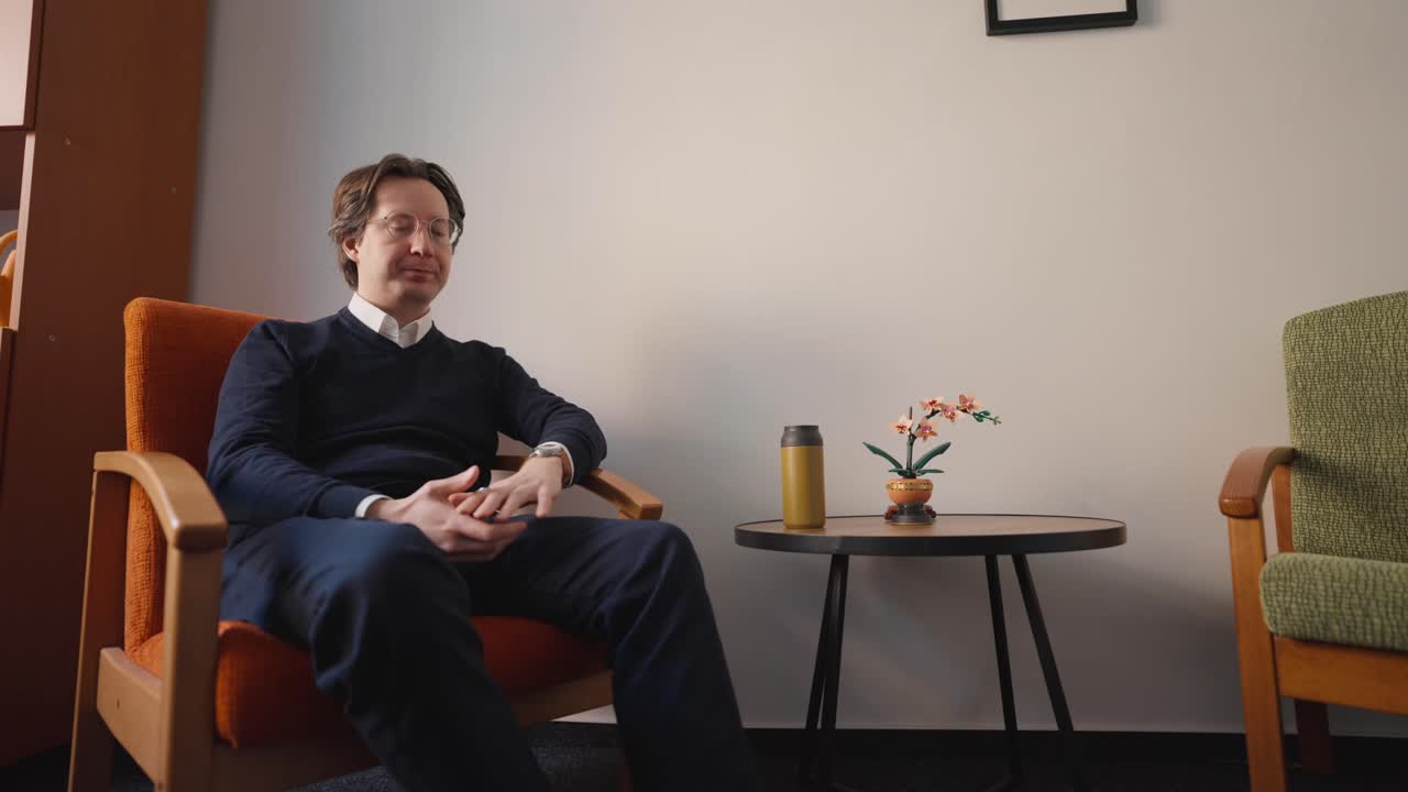 Sandalyede bekleyen, yorgunluk ve stres belirtileri gösteren bir adam. Evde saatinden zamanı kontrol ediyor.