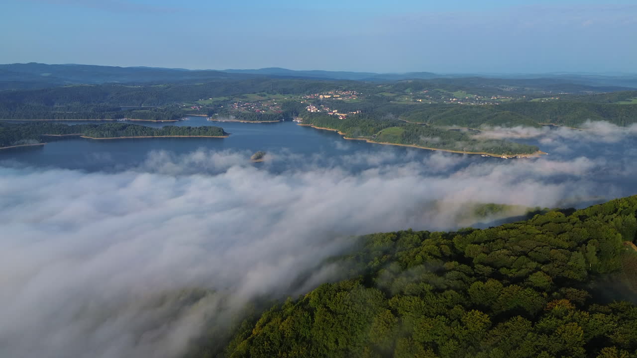 Aerial establishing shot of thick morning fog drifting across a calm lake, dense forest and distant village under clear blue sky. Solina, Poland
