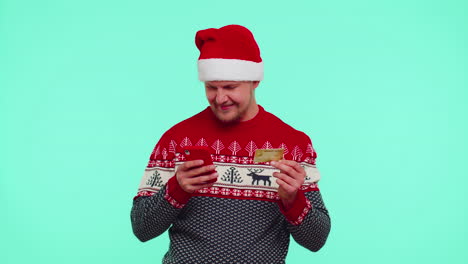 Man-in-Christmas-sweater-using-credit-bank-card,-smartphone-while-transferring-money-shopping-online