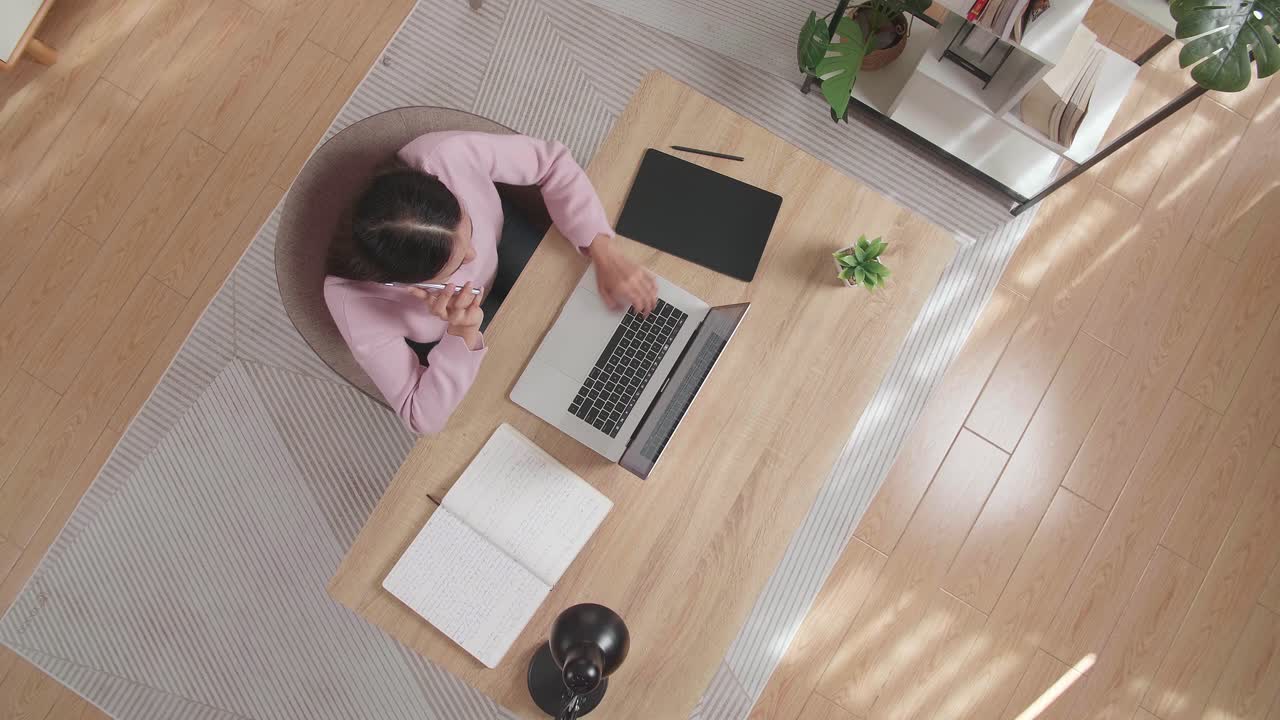 Top View Zoom Out Shot Of A Woman Talking On Smartphone While Sitting At The Wooden Desk Typing On A Laptop At Home