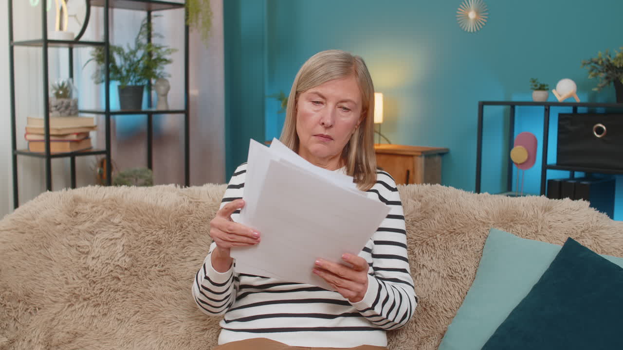 Mature woman reviewing documents and bills on sofa expressing uncertainty and frustration at home