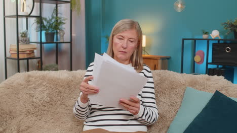 mature woman reviewing documents and bills on sofa expressing uncertainty and frustration at home