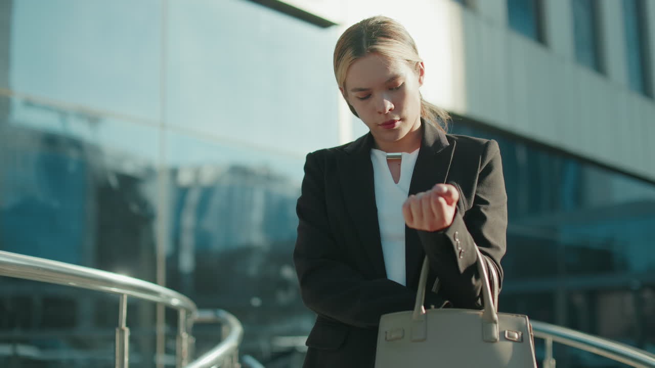 Female lecturer preparing for lecture, standing outside modern glass building, checking handbag to retrieve car keys, focused expression, urban reflections visible on structure
