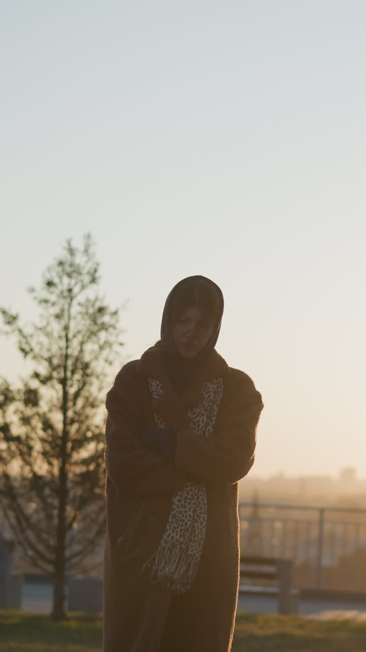 눈물을 흘리는 소녀, 팔에 손을 접고, 코트와 스카프를 입고, 해가 지는 동안 공원에서 돌로 된 기념비 앞에 서 있습니다. 그녀의 머리카락은 부분적으로 얼굴을 고, 그녀의 슬픈 분위기를 반영합니다.