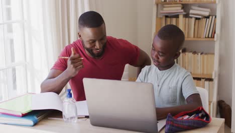 Afroamerikanischer-Vater-Hilft-Seinem-Sohn-Bei-Den-Hausaufgaben,-Während-Er-Mit-Laptop-Und-Desinfektionsflasche-Sitzt