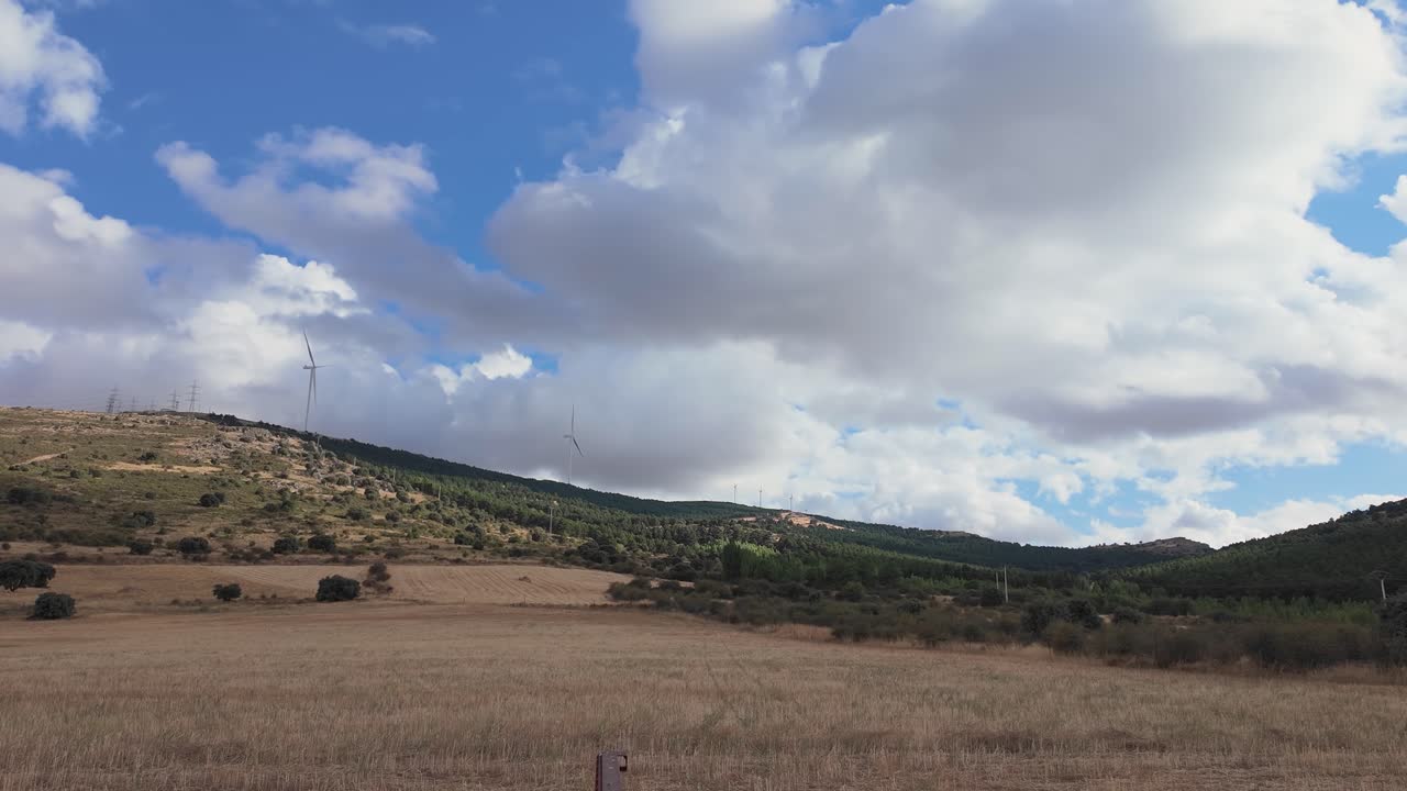 Un timelapse statico cattura nuvole bianche intense che si muovono rapidamente su un paesaggio rurale a Segovia, con turbine eoliche che generano energia verde sulle colline in lontananza