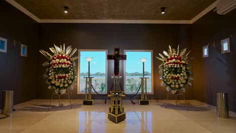 moving shot of the interior of a luxurious wake room with two flower arrangements, windows through which daylight enters and a cross in the background