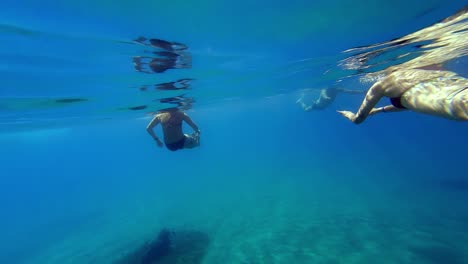 two women in black bikinis are swimming in a clear blue ocean with the sun shining down, with the camera submerged in the water just below the surface, and facing two women swimming away from it