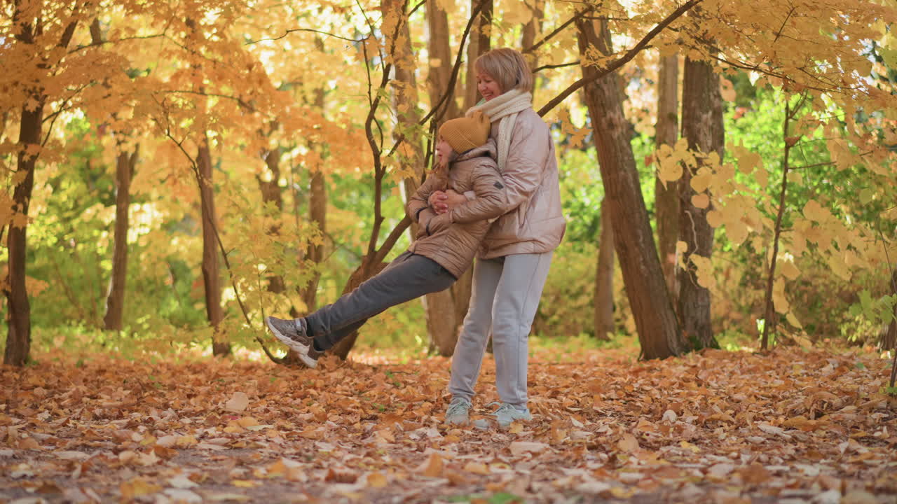 mother holding child from behind as they spin joyfully among golden autumn trees, child lifting legs with laughter while surrounded by colorful foliage and dry leaves covering forest ground
