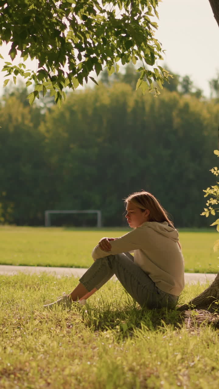 빛이 비치는 공원에서 나무 에 혼자 앉아있는 여성, 평온한 표정과 빛은 그녀를 둘러싼 부드러운 그림자를 던집니다. 배경에는 울창한 초록색, 나무, 멀리 있는 축구 골포스트가 있습니다.