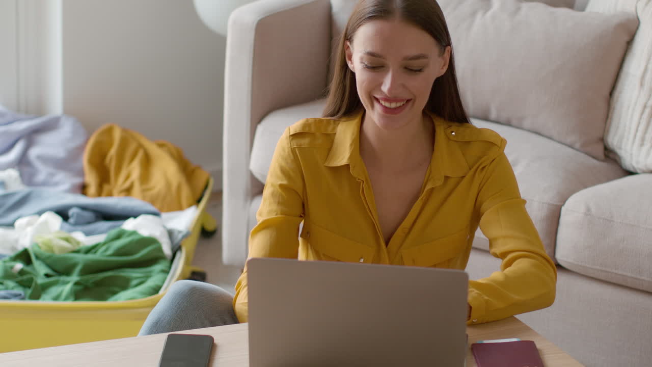 Woman working on laptop while packing for a trip at home