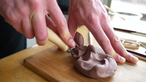 close up on the hands of an artist carving and cutting brown modeling clay with a metal tool before sculpting and shaping the clay with his fingers slide right