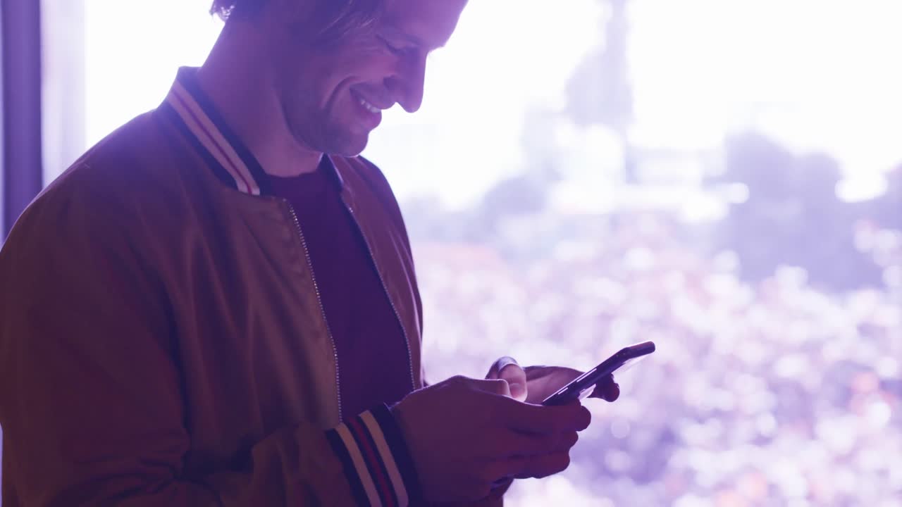 Caucasian male smiling and using his phone in a street in an evening,bright lights in the background