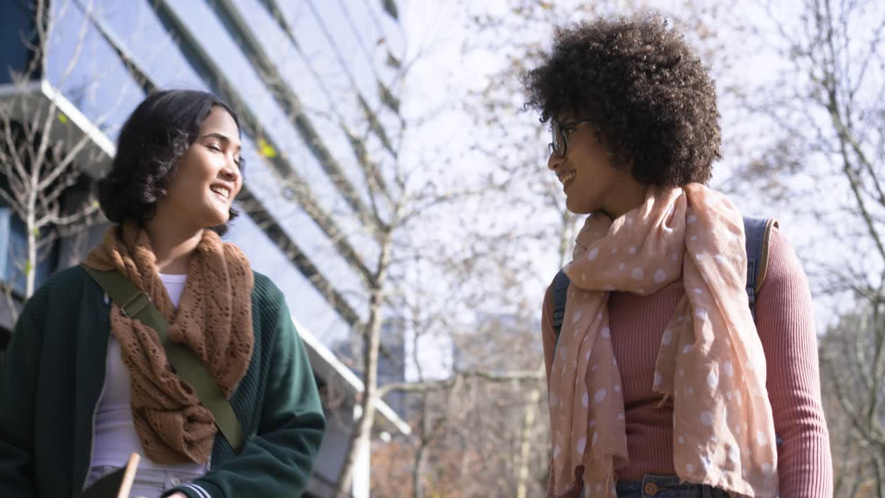 Diverse female friends walking path by glass building, chatting and gesturing with scarves and bags